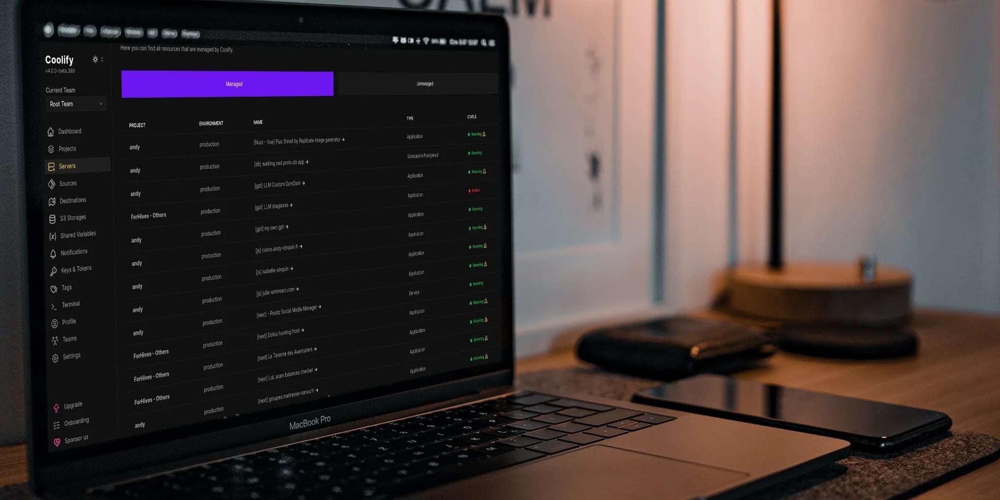 Close-up view of a MacBook Pro displaying the Coolify project management tool interface, with project details and status indicators, surrounded by smartphones on a textured surface.