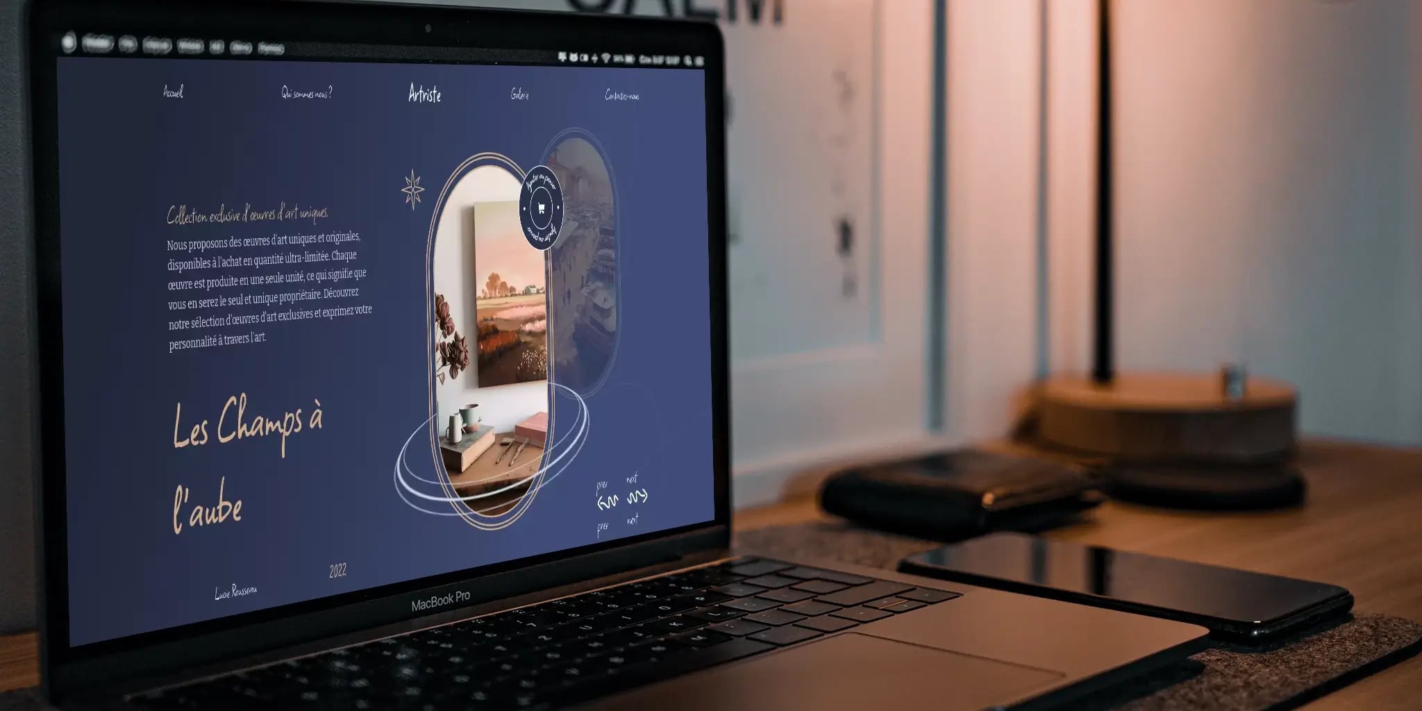 Close-up of MacBook Pro screen showing webpage in French with artistic imagery, smartphone and graphics tablet on wooden table in workspace setting