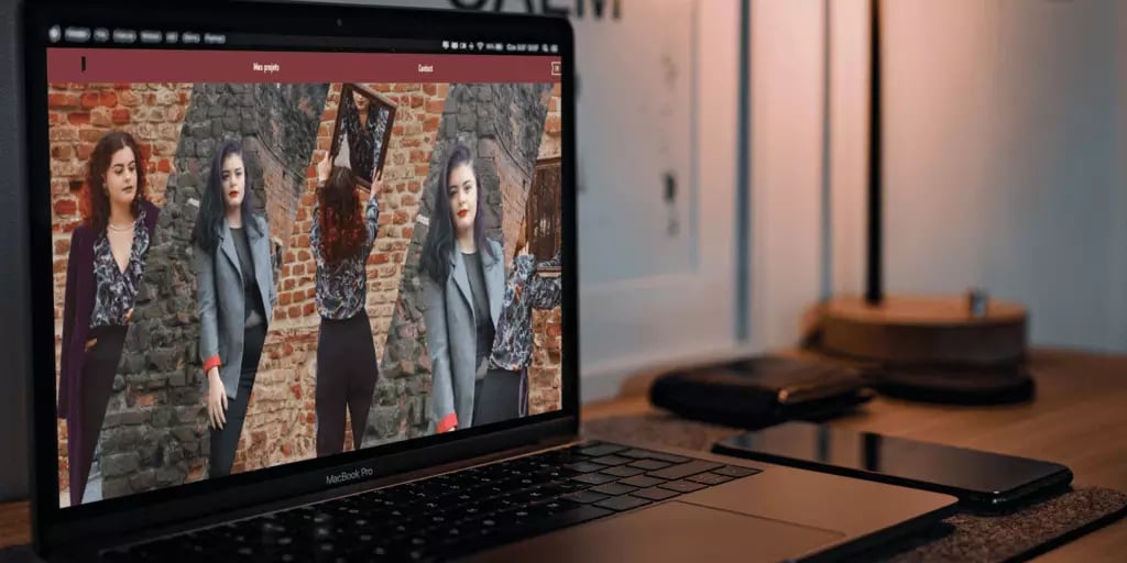 Laptop on a wooden table displaying images of a woman in different poses next to a brick wall with a smartphone and closed notebook nearby