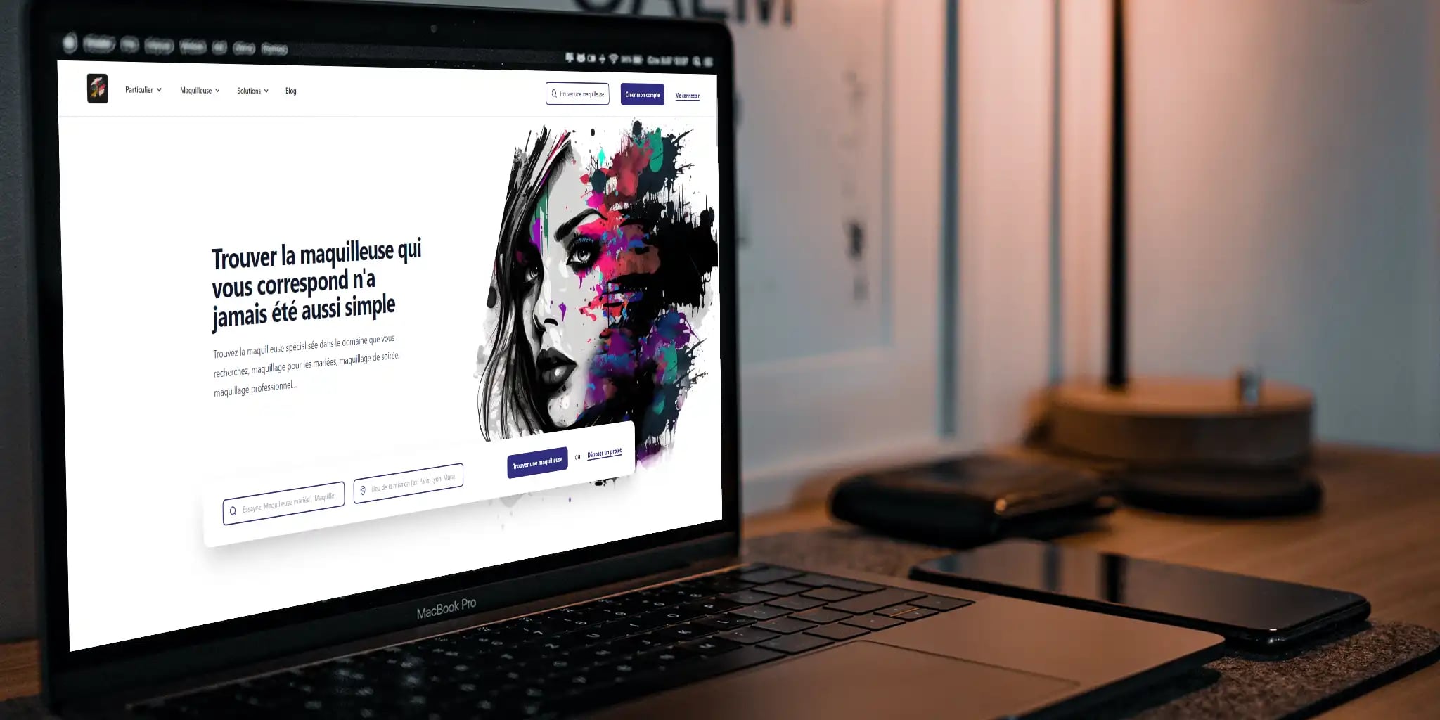 An open laptop displaying a French makeup artist website with colorful paint splashes on a woman's face, surrounded by a smartphone, tablet, and desk lamp on the desk