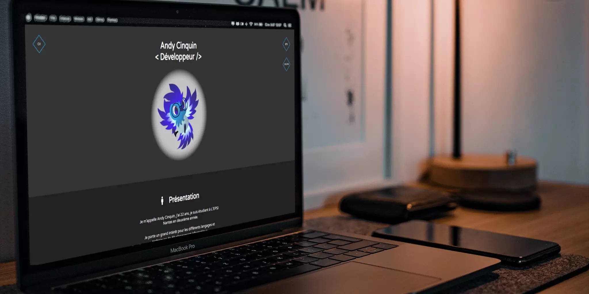A laptop on a wooden desk displaying a personal portfolio website, with a smartphone and a desk lamp with warm light in a modern workspace setting.