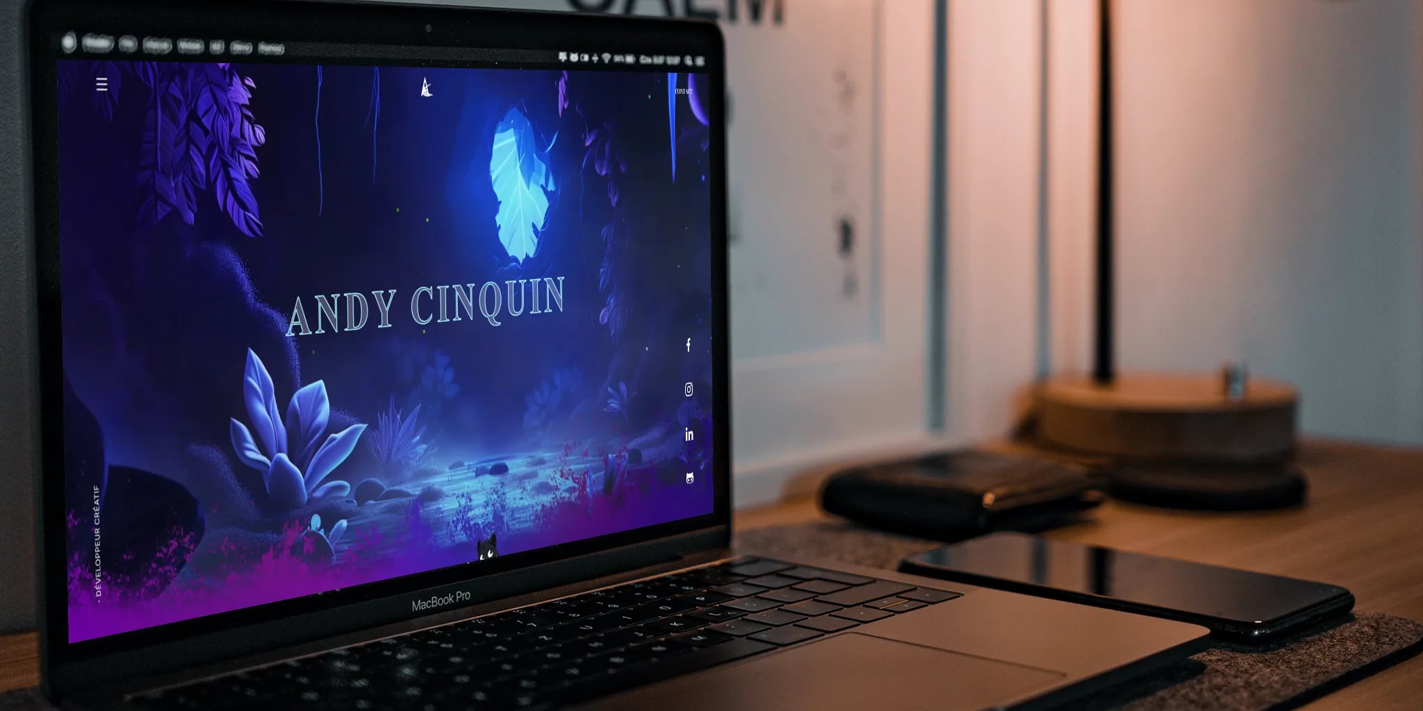 Laptop displaying personal branding 'Andy Cinquin' with artistic graphics, accompanied by smartphone, tablet with stylus, and other desk items in softly lit room