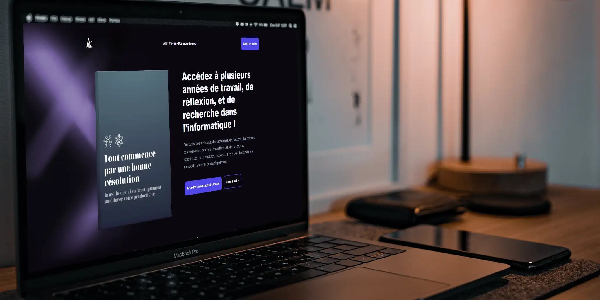 Close-up view of a laptop displaying a webpage with French text on a desk with a smartphone, tablet, notebook, and desk lamp in a modern workspace setting